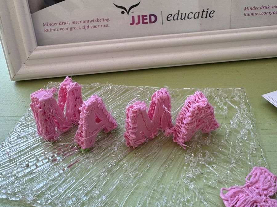 Het woord mama, gemaakt van roze 3D-printdraad, staat op een glasplaat voor een ingelijste JJED-poster.